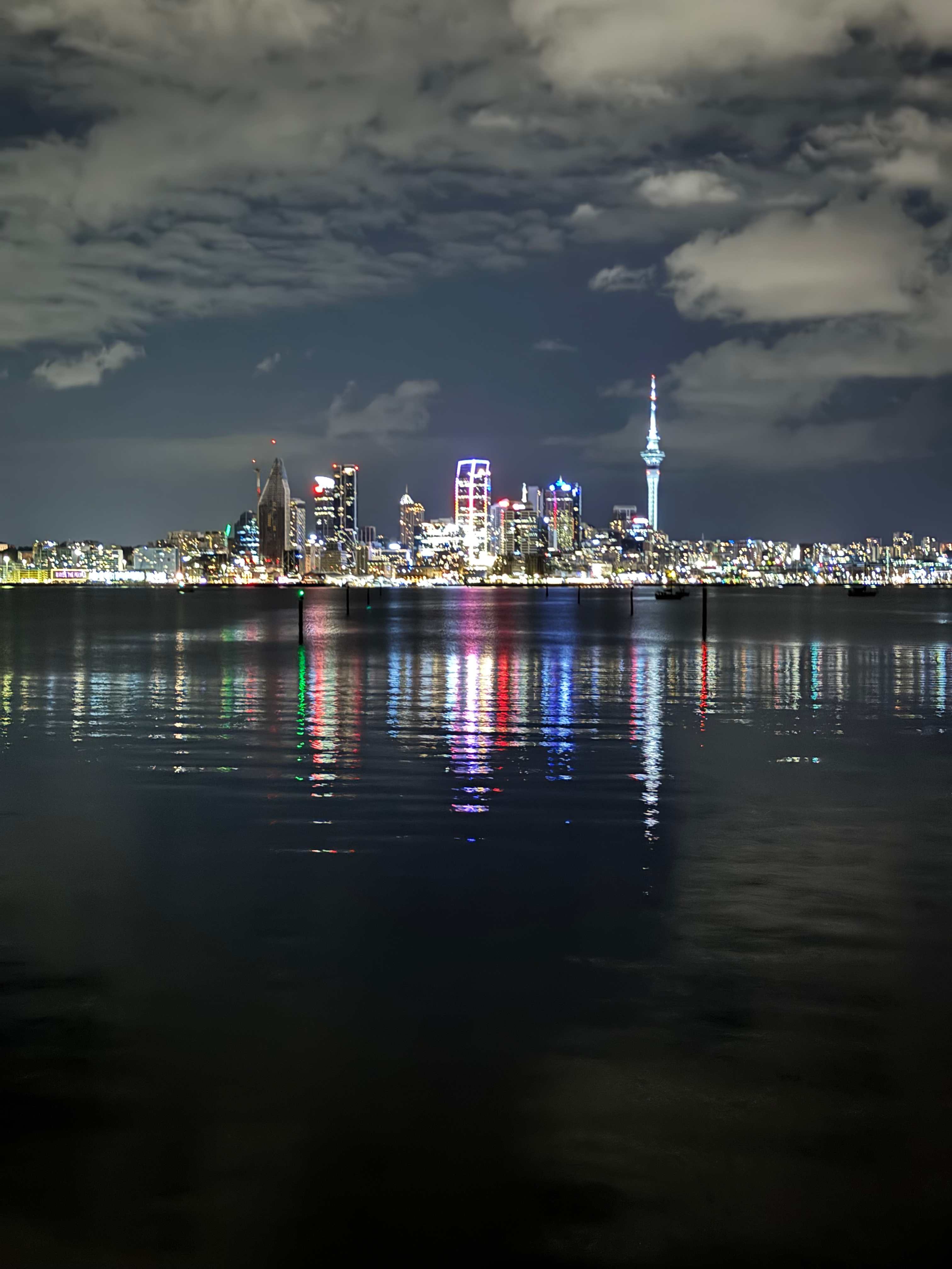 Auckland city skyline at night with the iconic Sky Tower illuminated and colorful lights reflecting on the harbor waters