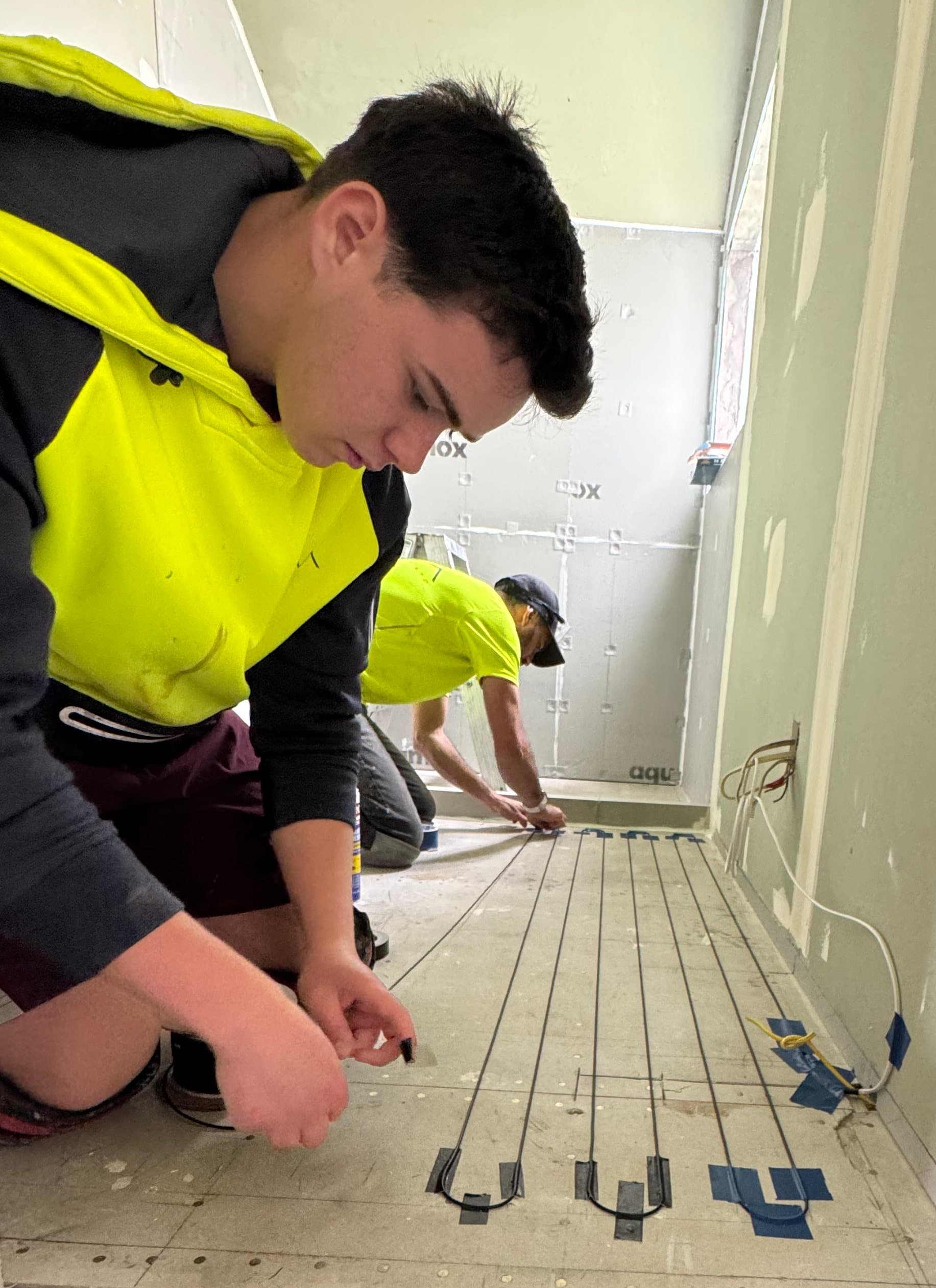 Professional installers in high-visibility clothing laying electric undertile heating cables on a bathroom floor, securing them with blue tape before tiling