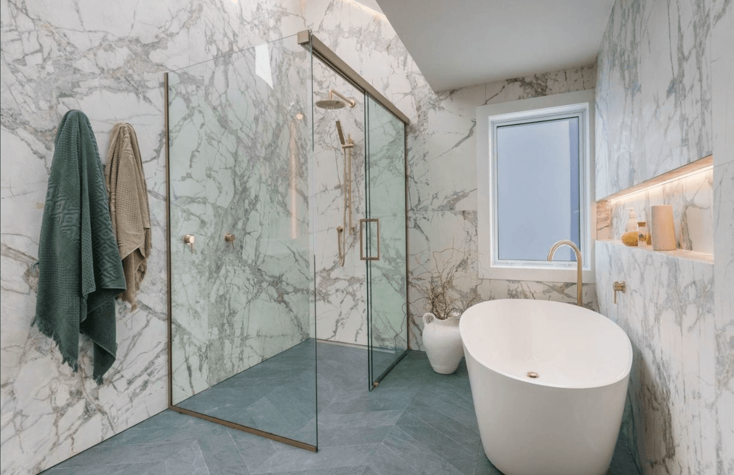 Elegant bathroom with white marble walls, gold-framed glass shower, freestanding white tub, and grey floor tiles