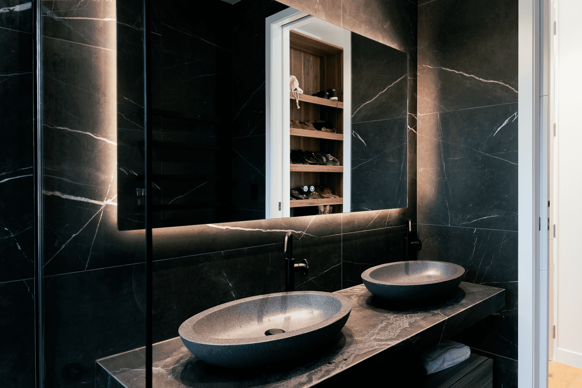 Sophisticated ensuite with black marble walls, dual vessel sinks, backlit mirror with shoe storage, and ambient lighting