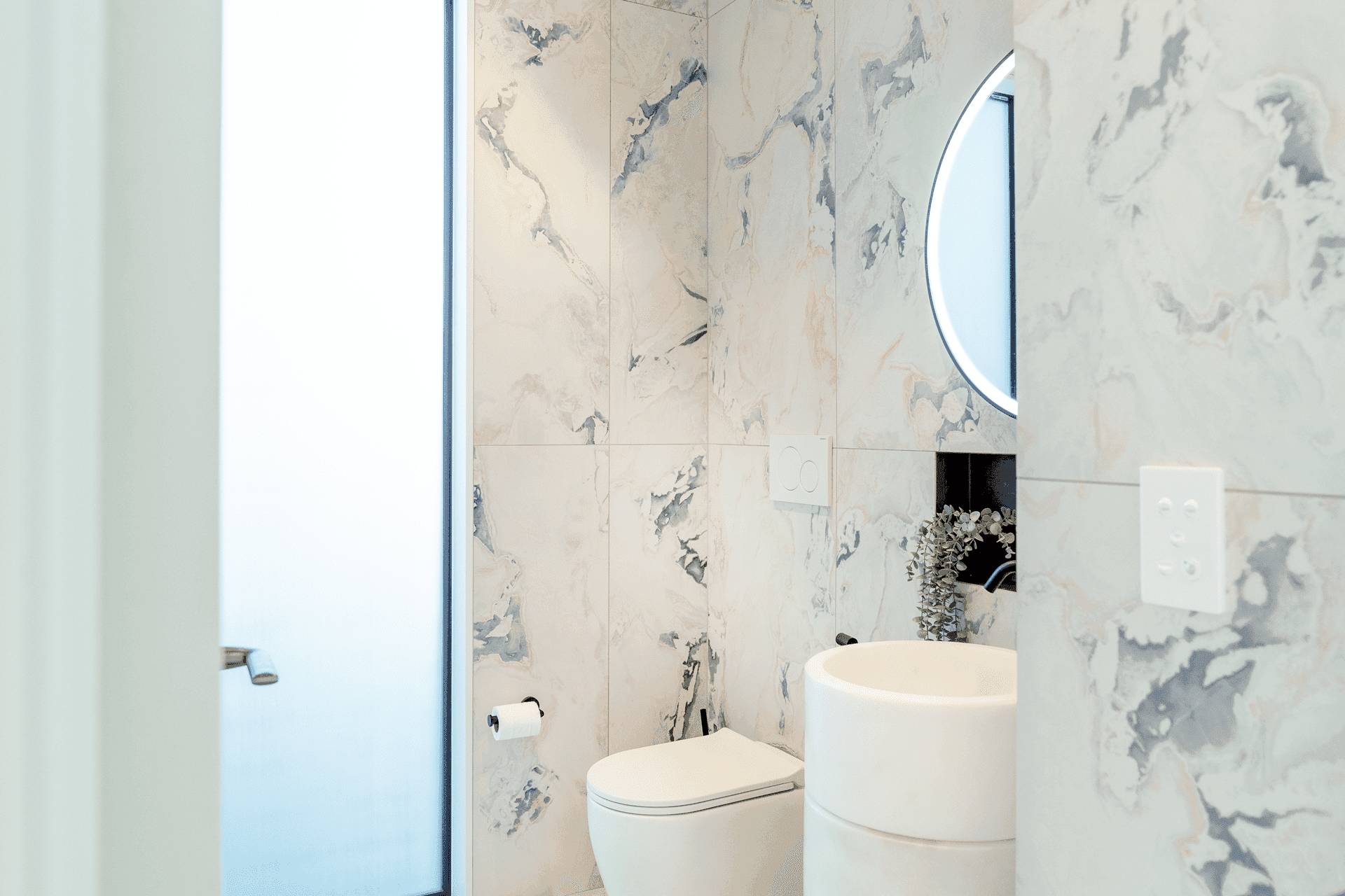 Minimalist powder room with artistic marble walls, round mirror, and cylindrical pedestal sink