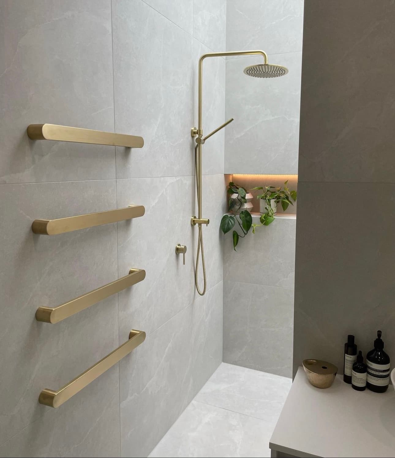 Minimalist bathroom with light marble tiles, brushed gold towel rails, gold shower fixtures, and built-in niche with plants