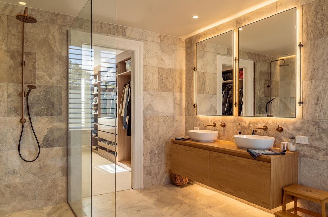 Modern bathroom with natural stone tiling, walk-in shower, double vessel sinks on wooden vanity, and backlit mirrors