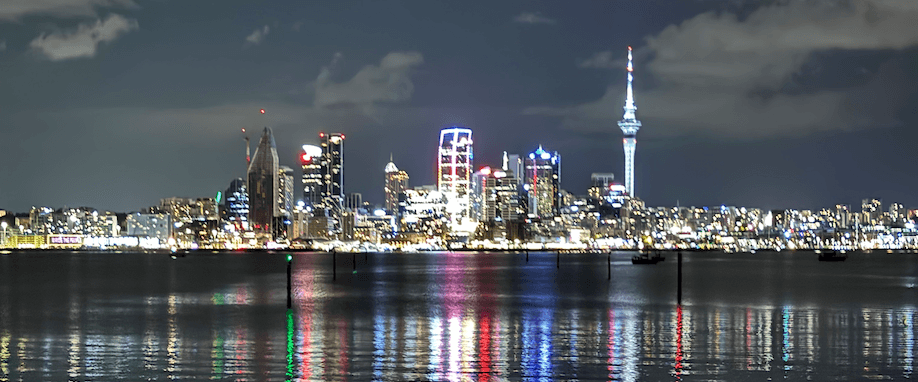 Auckland city skyline at night with the iconic Sky Tower illuminated and colorful lights reflecting on the harbor waters